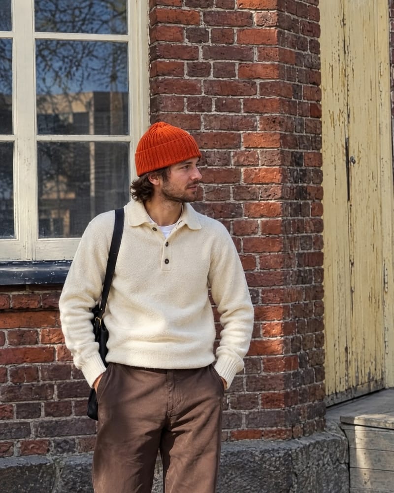Cream polo-collar knit and brown pants with an orange beanie, leaning on a brick wall